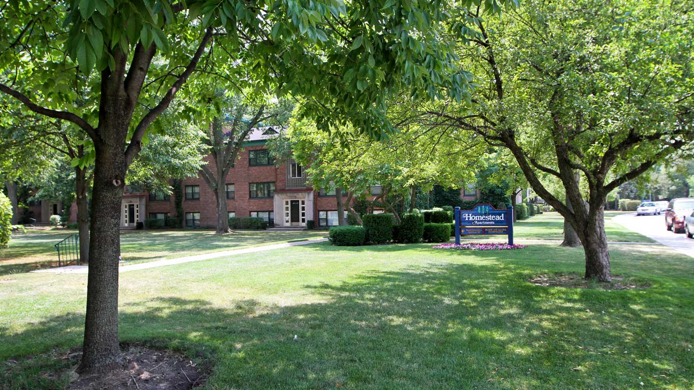 A brick apartment building partially obscured by trees, with a green lawn in the foreground and a sign reading "Homestead Apartments" near the sidewalk.