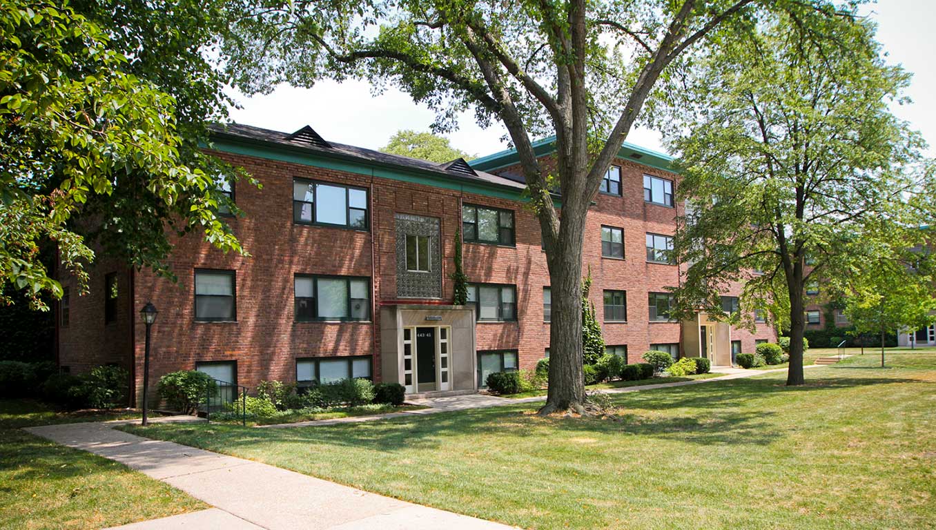 Three-story brick apartment building with green roof accents, large windows, and trees and greenery in the foreground. A concrete walkway leads up to the entrance.