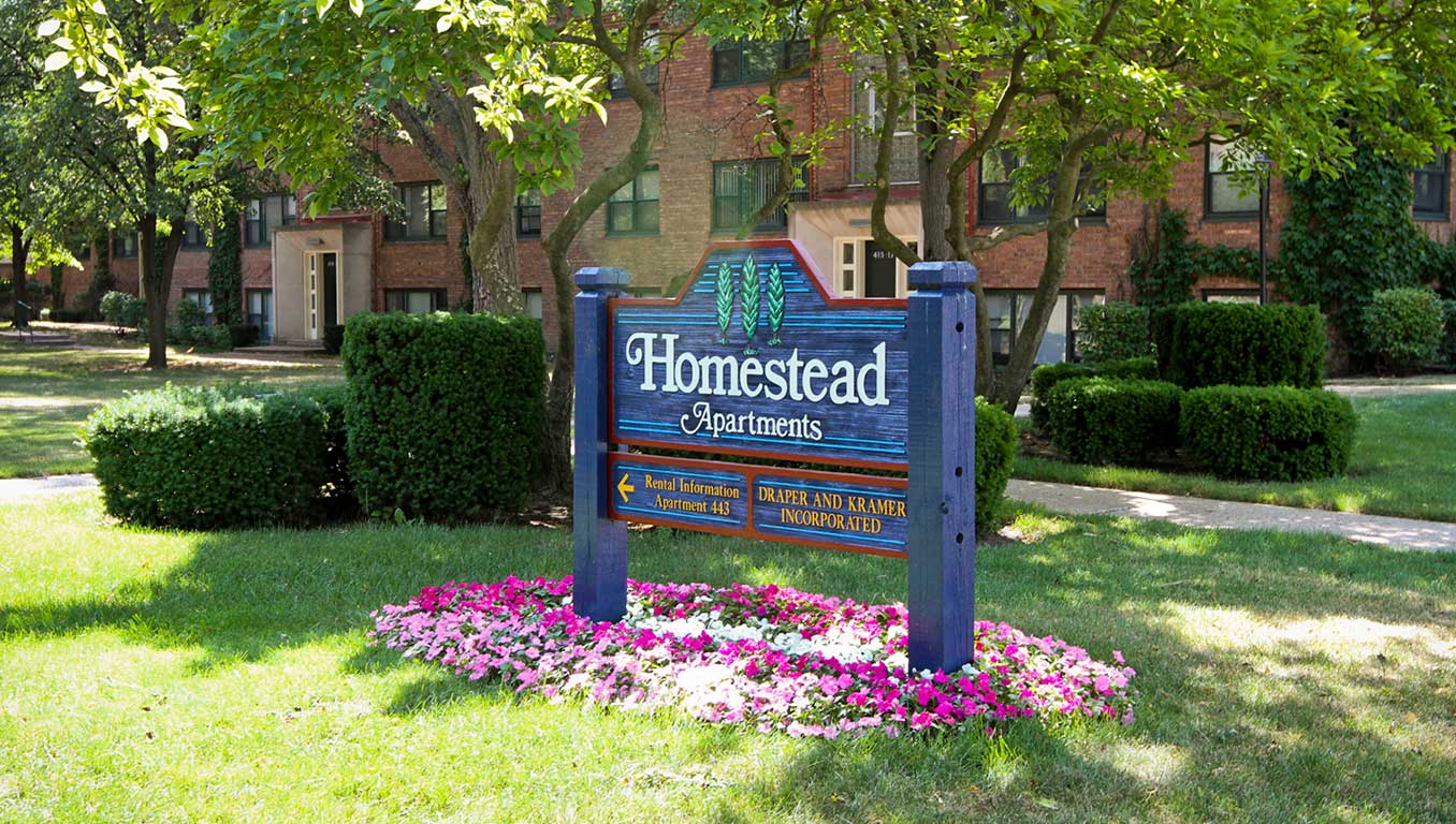 A wooden sign for Homestead Apartments stands on a lawn of flowers and green grass, with brick apartment buildings and trees in the background.