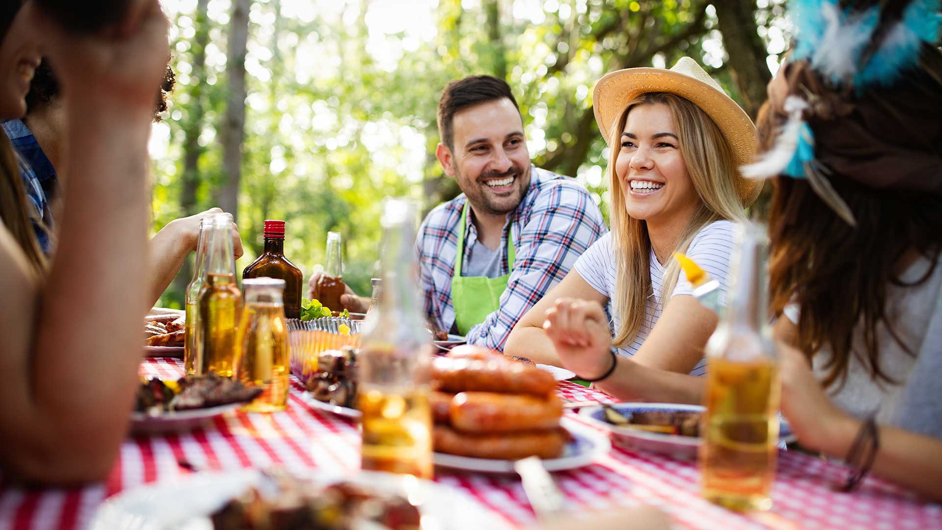 A group of people enjoying a picnic at a table outdoors. Plates of food and bottles are visible on the red checkered tablecloth. Trees are in the background.