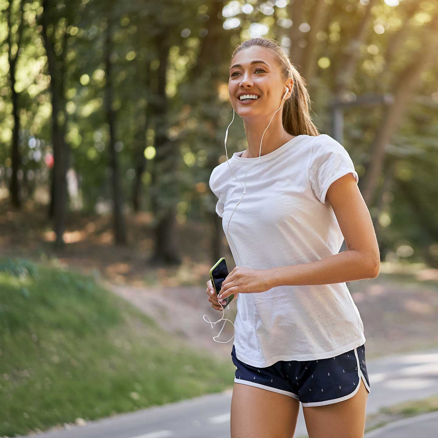 A woman jogs on a path in a park, wearing a white t-shirt, blue shorts, and earphones, and holding a smartphone. She is smiling and looks focused. Trees and greenery are in the background.