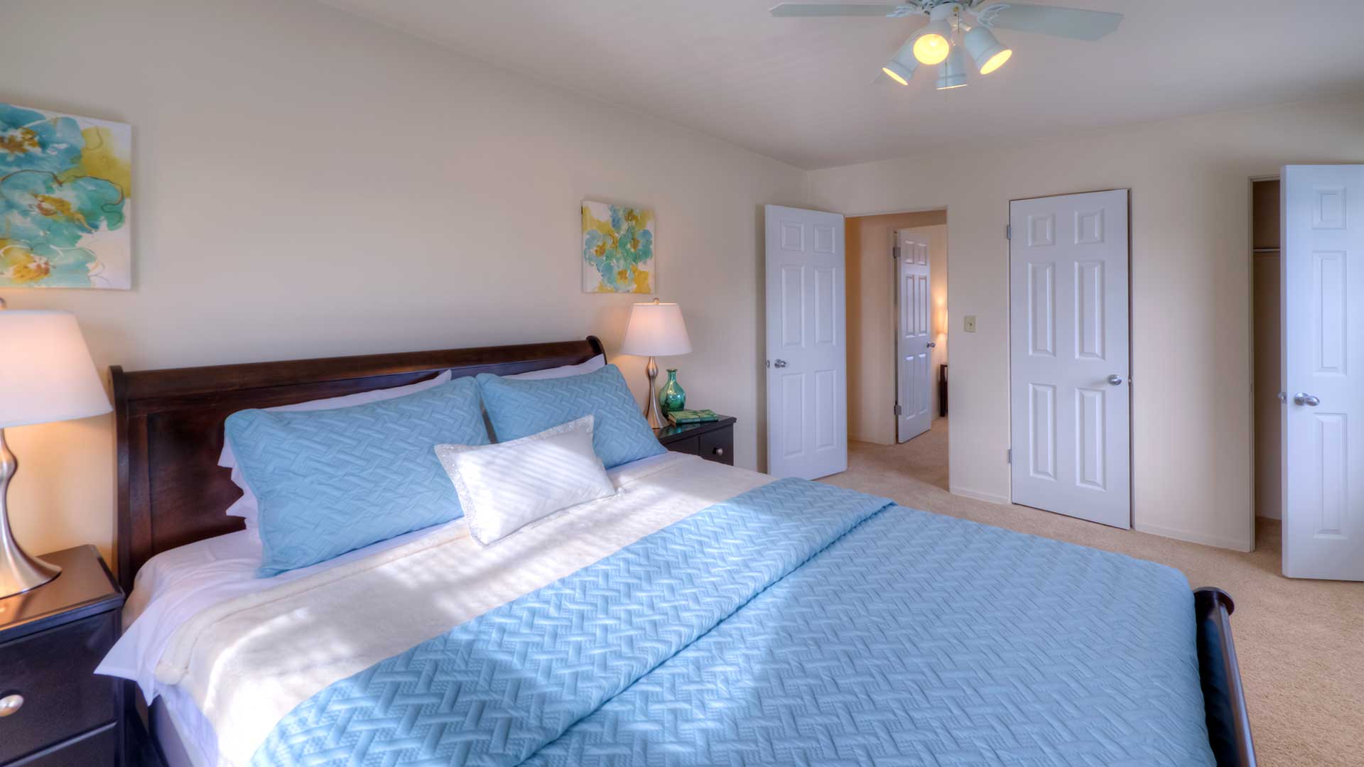 A bedroom with a blue bedspread, two open closet doors, a ceiling fan, a bedside lamp, and a painting on the wall.