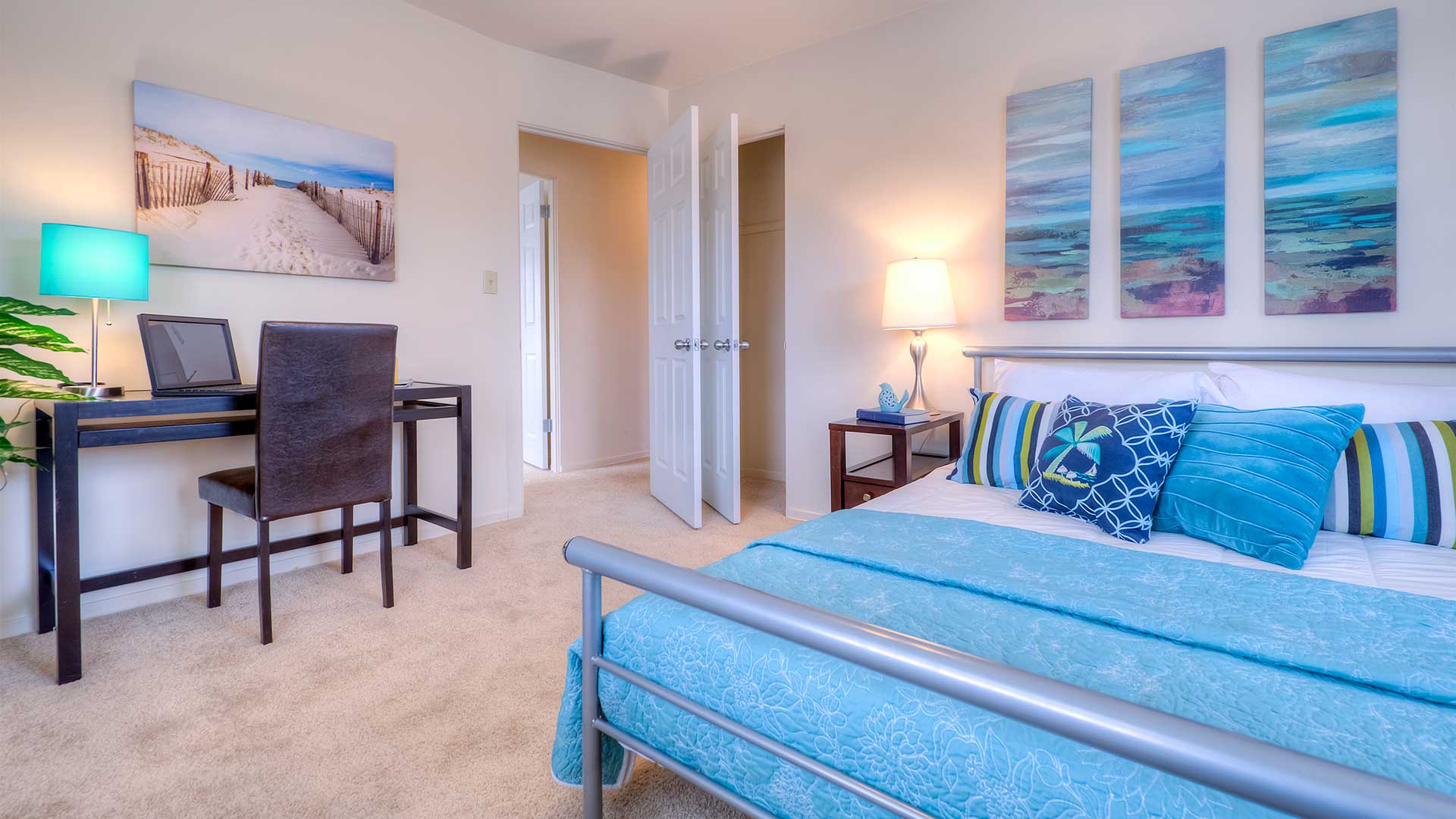 A tidy bedroom with a bed, blue linens, decorative pillows, two nightstands, a lamp, framed artwork, a desk, and an open door to a closet.