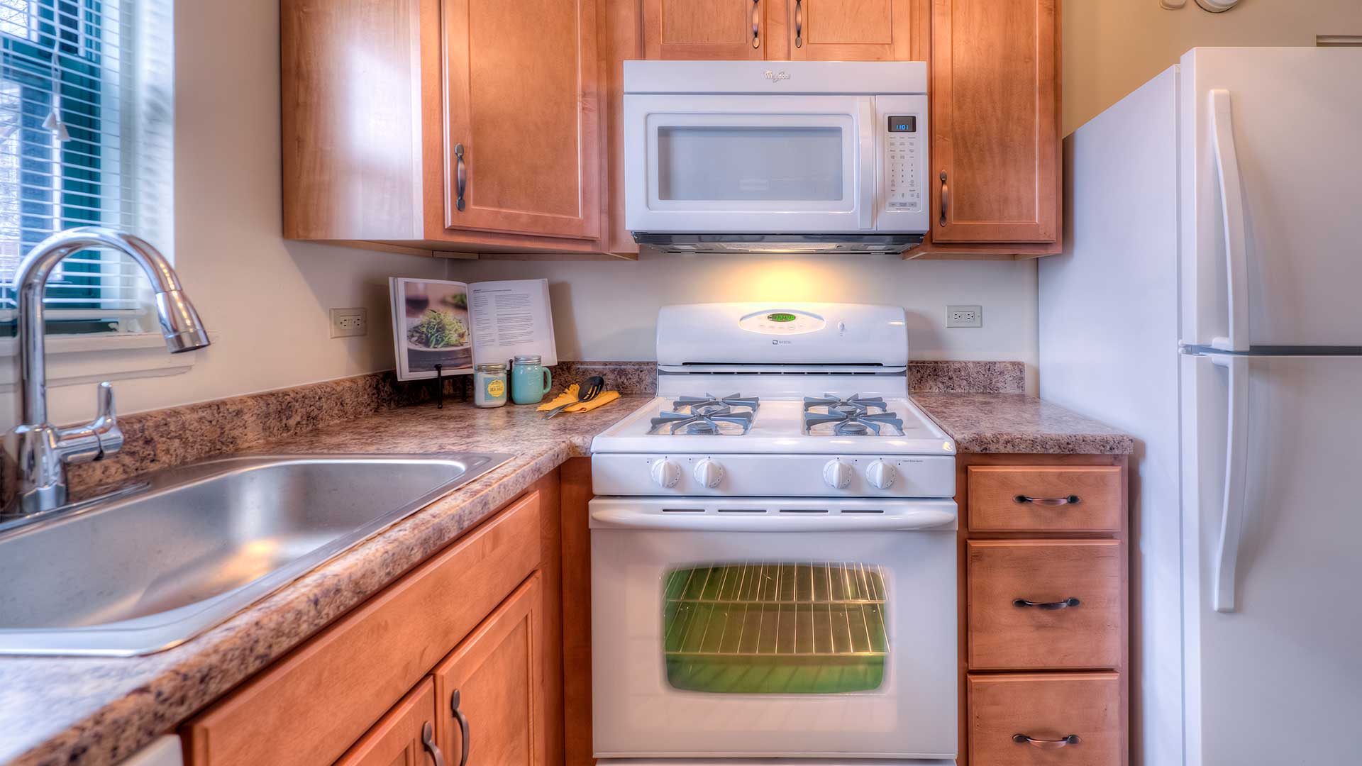 A kitchen with wooden cabinets, a gas stove, a microwave above it, a refrigerator, a sink with a faucet, and a granite countertop. Kitchen items are placed on the counter near the stove.