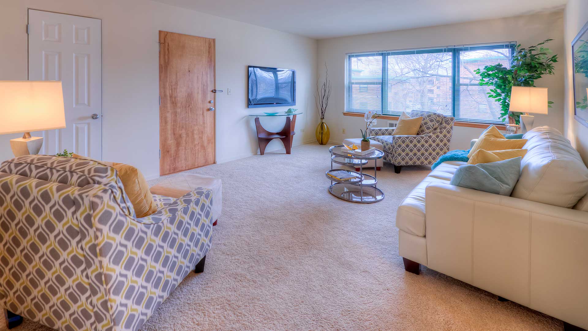 Bright living room with a white sofa, patterned armchairs, round glass coffee table, wall-mounted TV, wooden door, and large window. Room is decorated with lamps, plants, and various small decor pieces.