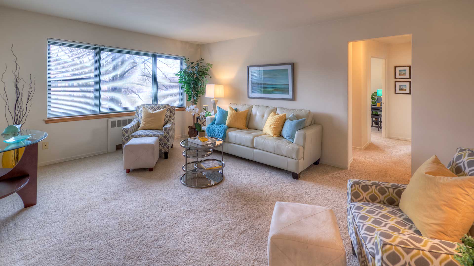 A cozy, well-lit living room with a white sofa adorned with yellow and teal pillows, two patterned armchairs, a glass coffee table, and a large window with blinds. A hallway is visible on the right.