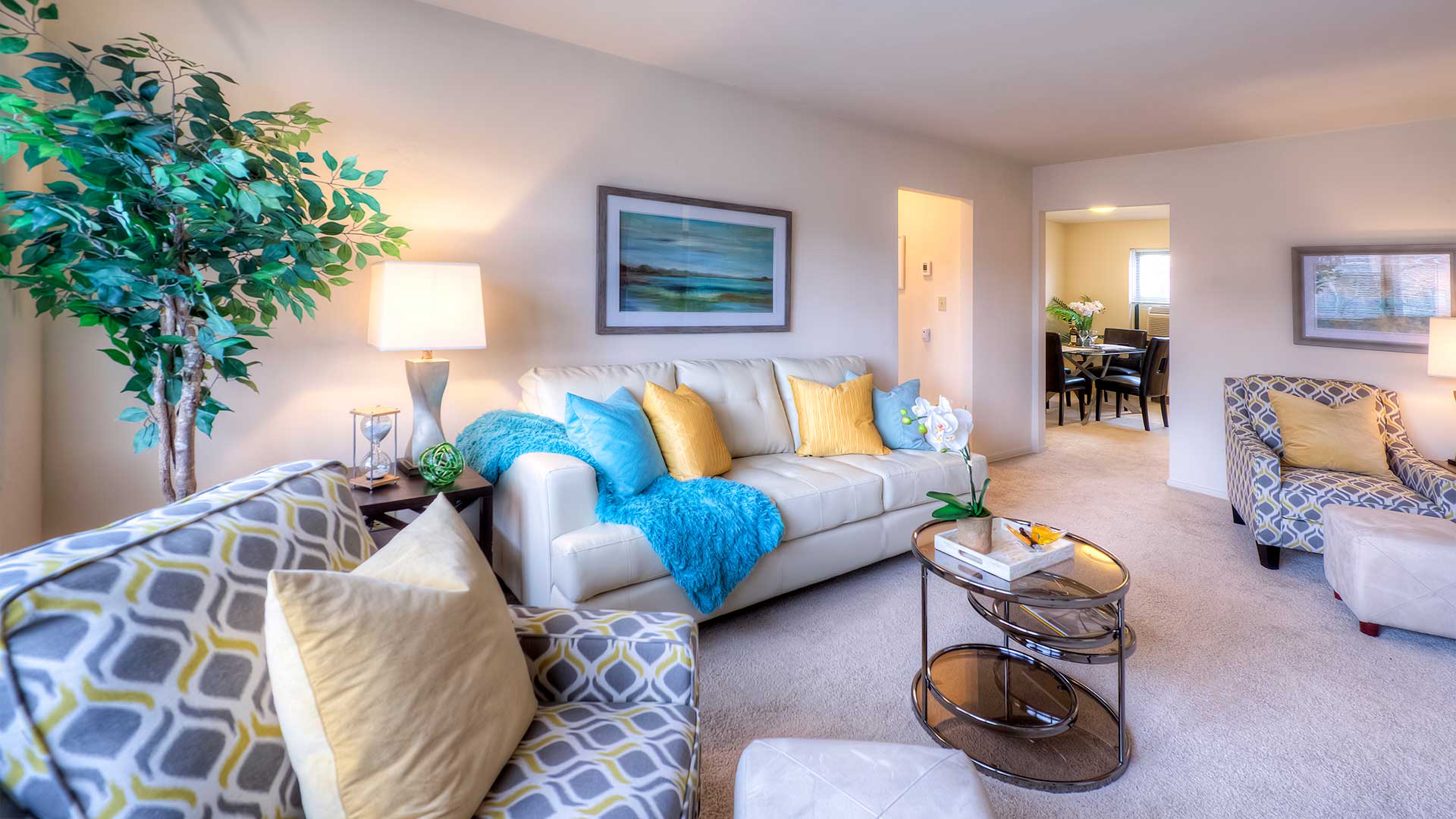 A cozy living room featuring a white couch with colorful pillows and a blue throw, patterned armchairs, a coffee table, and framed art on the walls. A dining area is visible in the adjoining room.